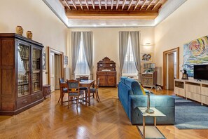 A living room with a wooden floor, a blue sofa, a dining table with chairs, a cabinet with glass doors, and a painting on the wall. - Domus Navona Apartment - Via dei Coronari (Rome)