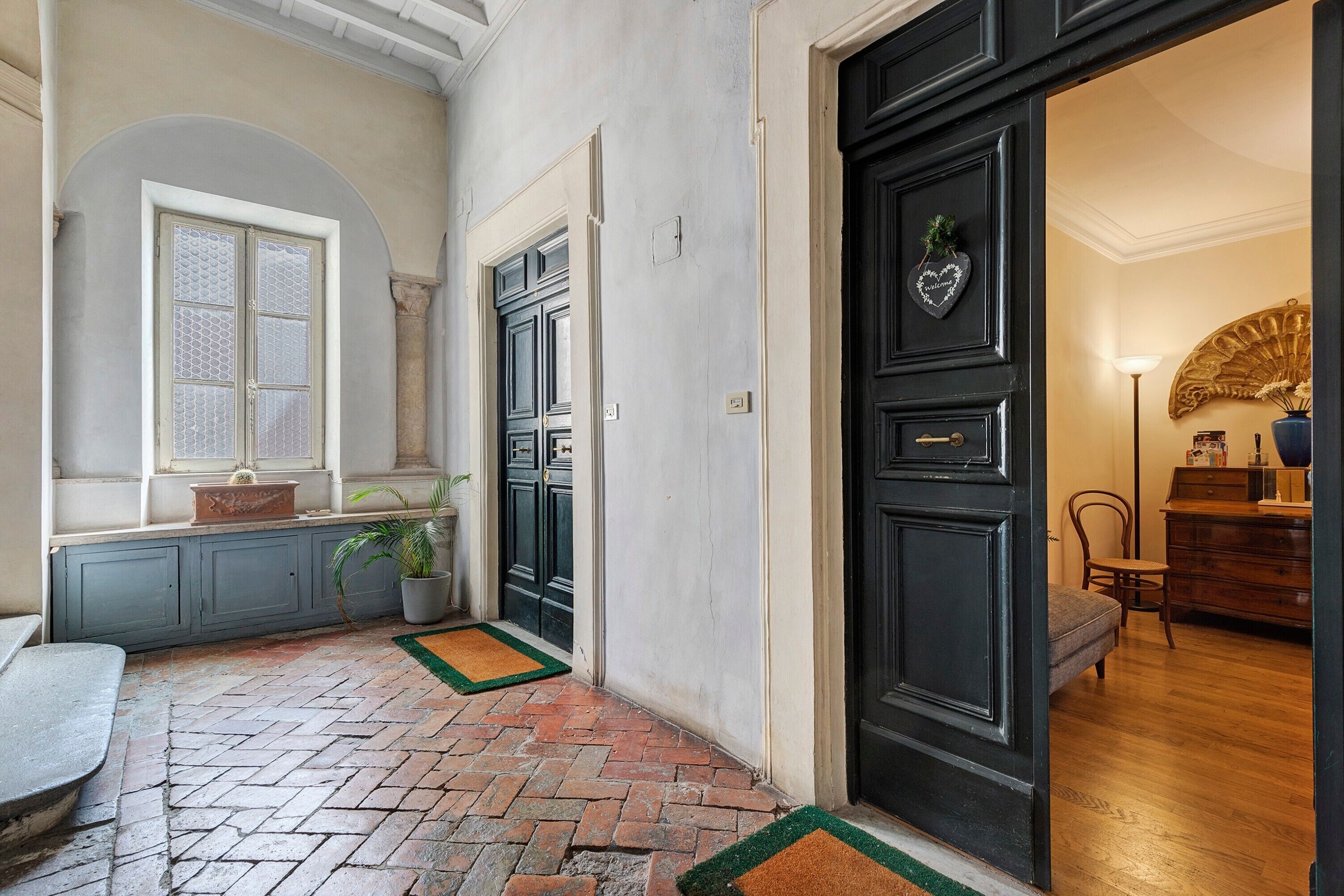 A hallway with a brick floor, a window with a view, a door with a heart-shaped welcome sign, and a chair in the room.