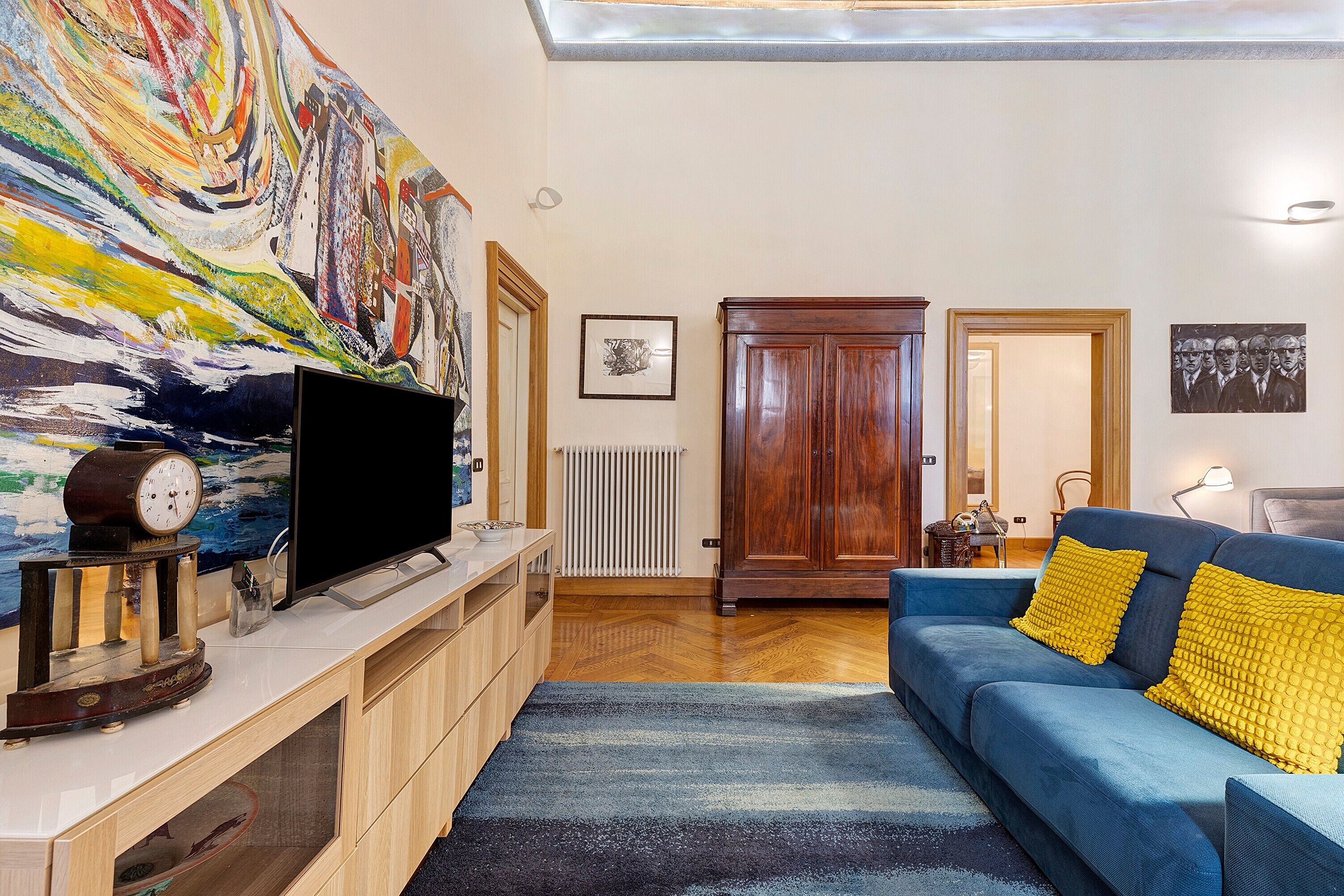 A living room with a flat-screen TV, wooden cabinets, a blue sofa with yellow cushions, a colorful abstract painting, and a wooden wardrobe.