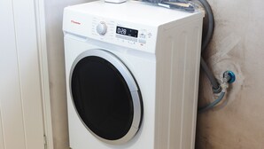 Máquina de lavar roupa branca com visor digital, tomada e mangueira ligada a um cano fixado na parede.