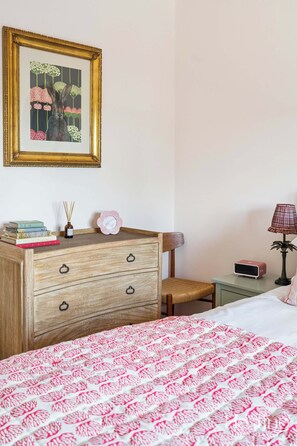 Un dormitorio con un aparador de madera, una silla, una mesita de noche con lámpara y una cama con colcha estampada.