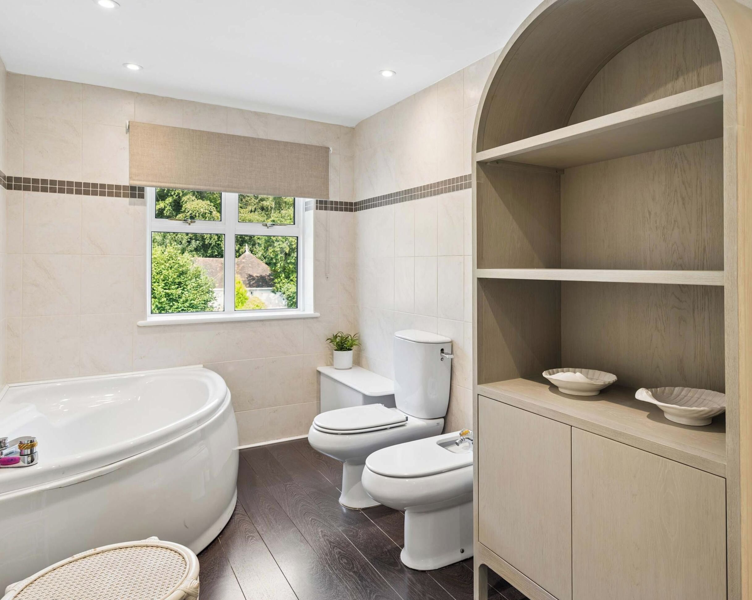 A bathroom with a bathtub, two toilets, a window with a view, and a shelf with bowls.
