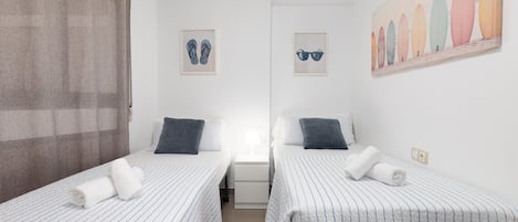 A room with two single beds, a nightstand, a lamp, and a painting of surfboards.