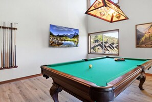 Une table de billard recouverte d’un drap vert, un porte-queues contenant des queues de billard, et deux tableaux encadrés représentant des paysages.