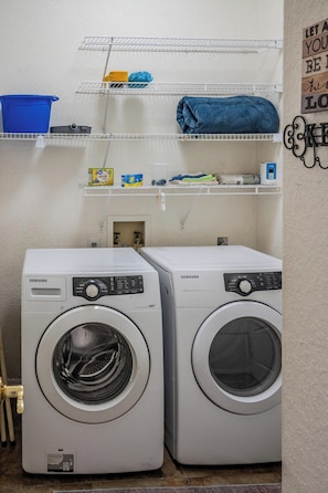 Two white Samsung front-loading washing machines in a laundry area with shelves above holding various items. - Cozy & Comfortable 3-Bed 2-Bath Sleeps 7 (Valrico)