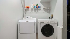 A laundry room with a front-loading washer and dryer, a shelf with cleaning supplies, and a vent hose. - The Weekender | Cozy Bridgeland Retreat | Sleeps 2 (Calgary)