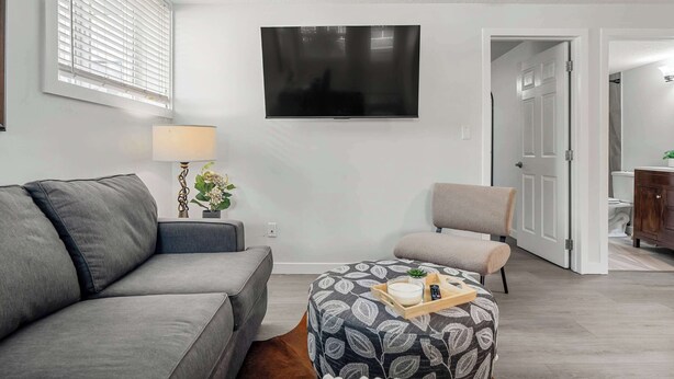 A modern living room with a grey sofa, a patterned ottoman, a television, and a door leading to a bathroom. - The Weekender | Cozy Bridgeland Retreat | Sleeps 2 (Calgary)