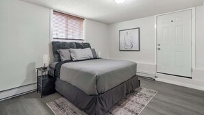 A bedroom with a bed, bedside table, nightstand, window with blinds, and a door. - The Weekender | Cozy Bridgeland Retreat | Sleeps 2 (Calgary)