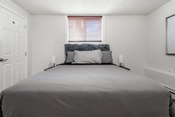 A bedroom with a bed, a window with blinds, a door, and a wall-mounted light fixture.