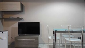 Een compacte kamer met een flatscreen televisie, een kleine koelkast en een tafel met glazen blad en witte stoelen.
