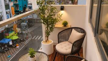 A balcony with wicker chairs and a small table, overlooking a city street with buildings and a clear sky.