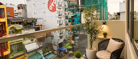 A balcony with a view of a cityscape, including a building with a red and white sign, a potted plant, and two wicker chairs.