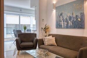 A modern living room with a brown sofa, a glass coffee table, and a cityscape painting on the wall. - Stylish 2BR in Sliema w/private balcony, WIFI & AC (Sliema)
