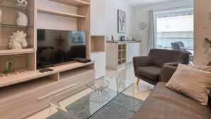 Uma sala de estar moderna com uma televisão de tela plana, uma mesa de centro de vidro, um sofá marrom e uma estante com objetos decorativos.