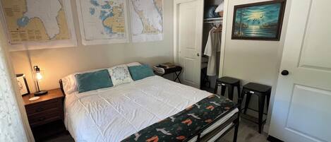 Una camera da letto con un letto, un comodino, una lampada, una mappa appesa al muro, un appendiabiti, una porta e un quadro.