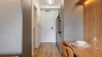 Um corredor estreito leva a uma área de refeições com uma mesa de madeira posta para dois, uma cadeira e uma taça de vinho. Uma porta com a placa 'SAO PAULO' é visível ao fundo.