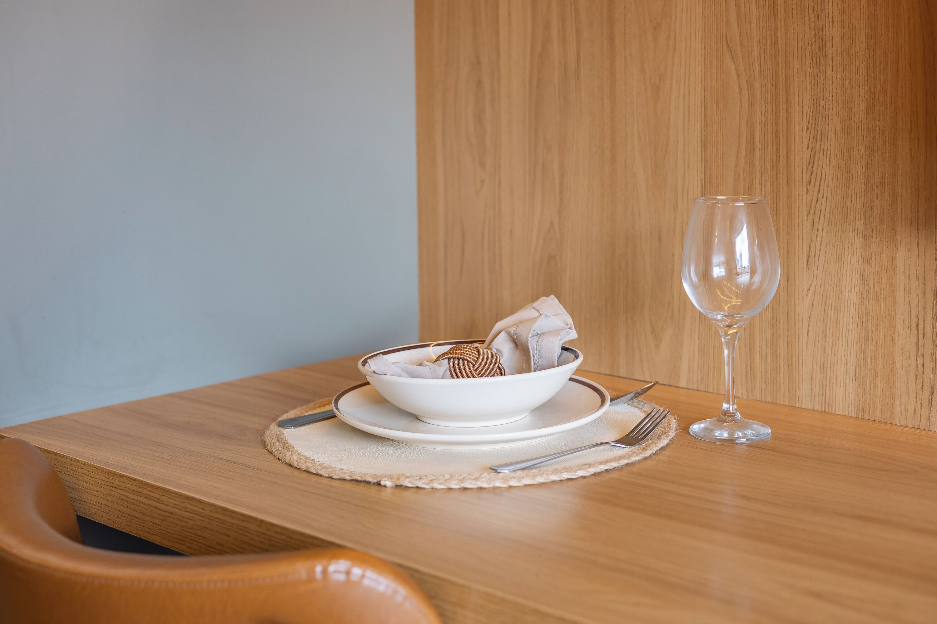 A wooden dining table with a white bowl, a folded napkin, a wine glass, and cutlery.