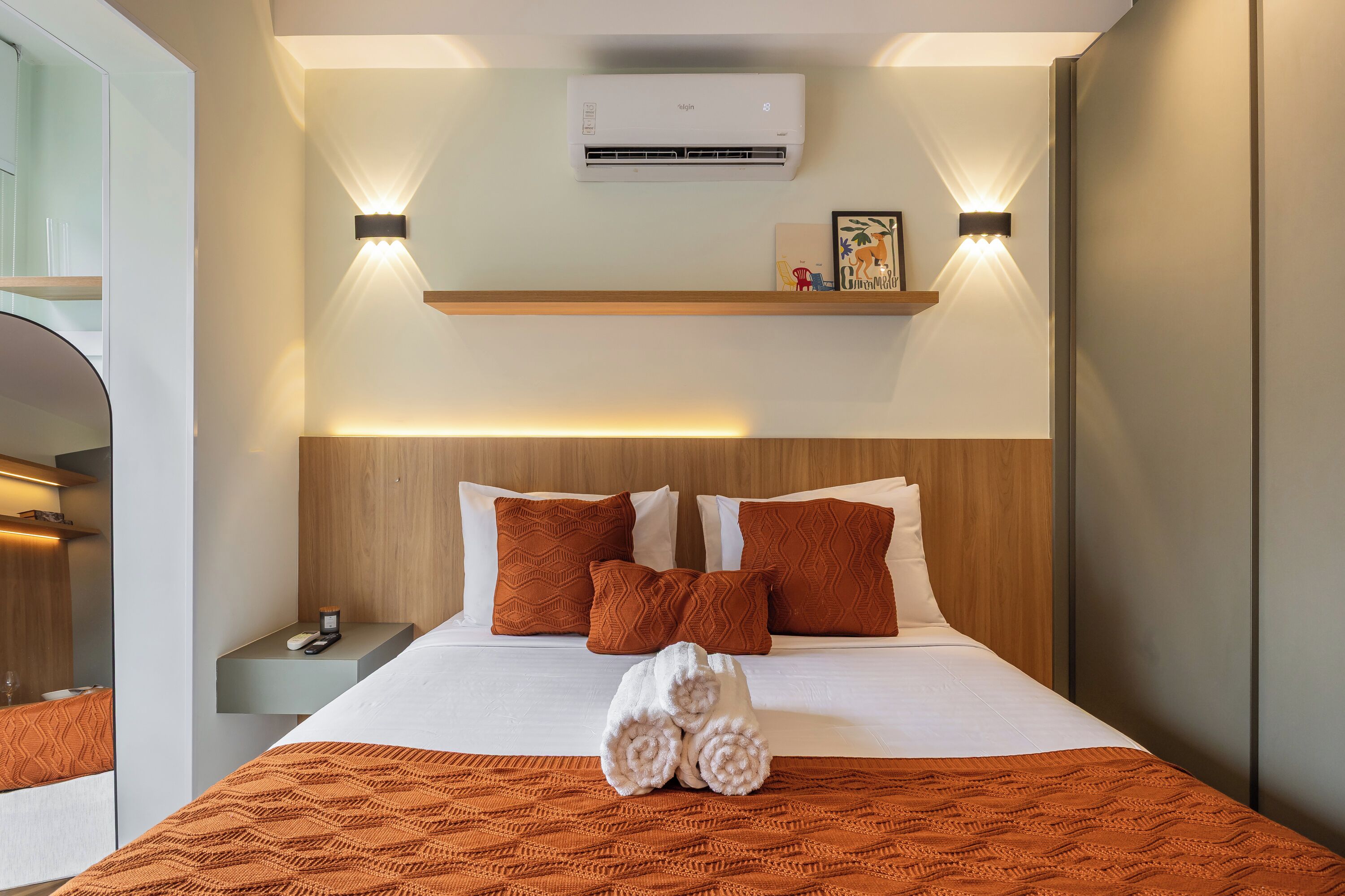 A hotel room with a bed, bedside tables, a nightstand, an air conditioning unit, wall-mounted lights, and a shelf with decorative items.