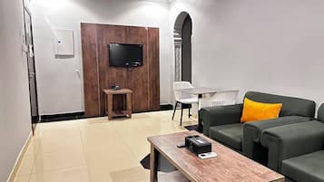 Un salon moderne avec un téléviseur à écran plat fixé sur un mur recouvert de panneaux en bois, un canapé agrémenté d’un coussin jaune, une table basse et une table à manger blanche avec des chaises.