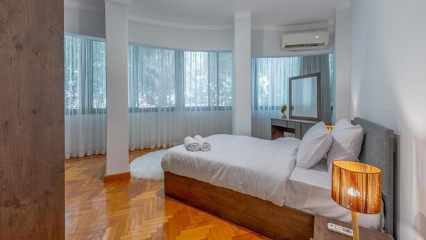 Une chambre moderne avec un plancher en bois, un lit recouvert de draps blancs, une lampe de chevet et une grande fenêtre avec des rideaux.