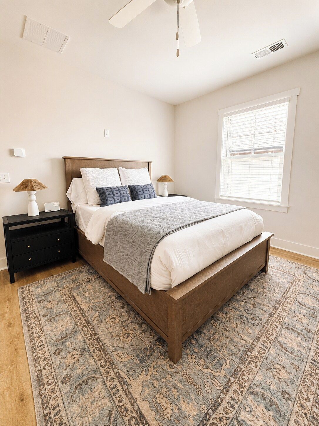 Une chambre à coucher comprenant un lit, des tables de chevet, un ventilateur de plafond, une fenêtre avec des stores et un tapis à motifs.