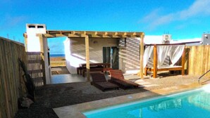Zona de piscina con terraza de madera, sillas de descanso y un área de estar cubierta.