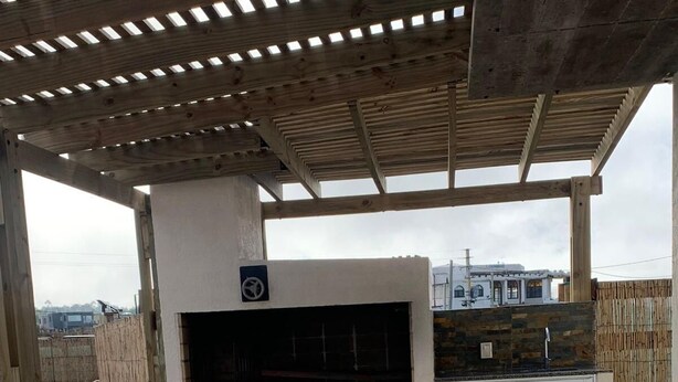 Un patio al aire libre con una mesa y bancos de madera, una parrilla de barbacoa empotrada y un muro de piedra al fondo.