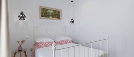 Un dormitorio con estructura de cama metálica blanca, una mesita de noche pequeña con cubierta de vidrio, un cuadro enmarcado en la pared y dos lámparas colgantes.