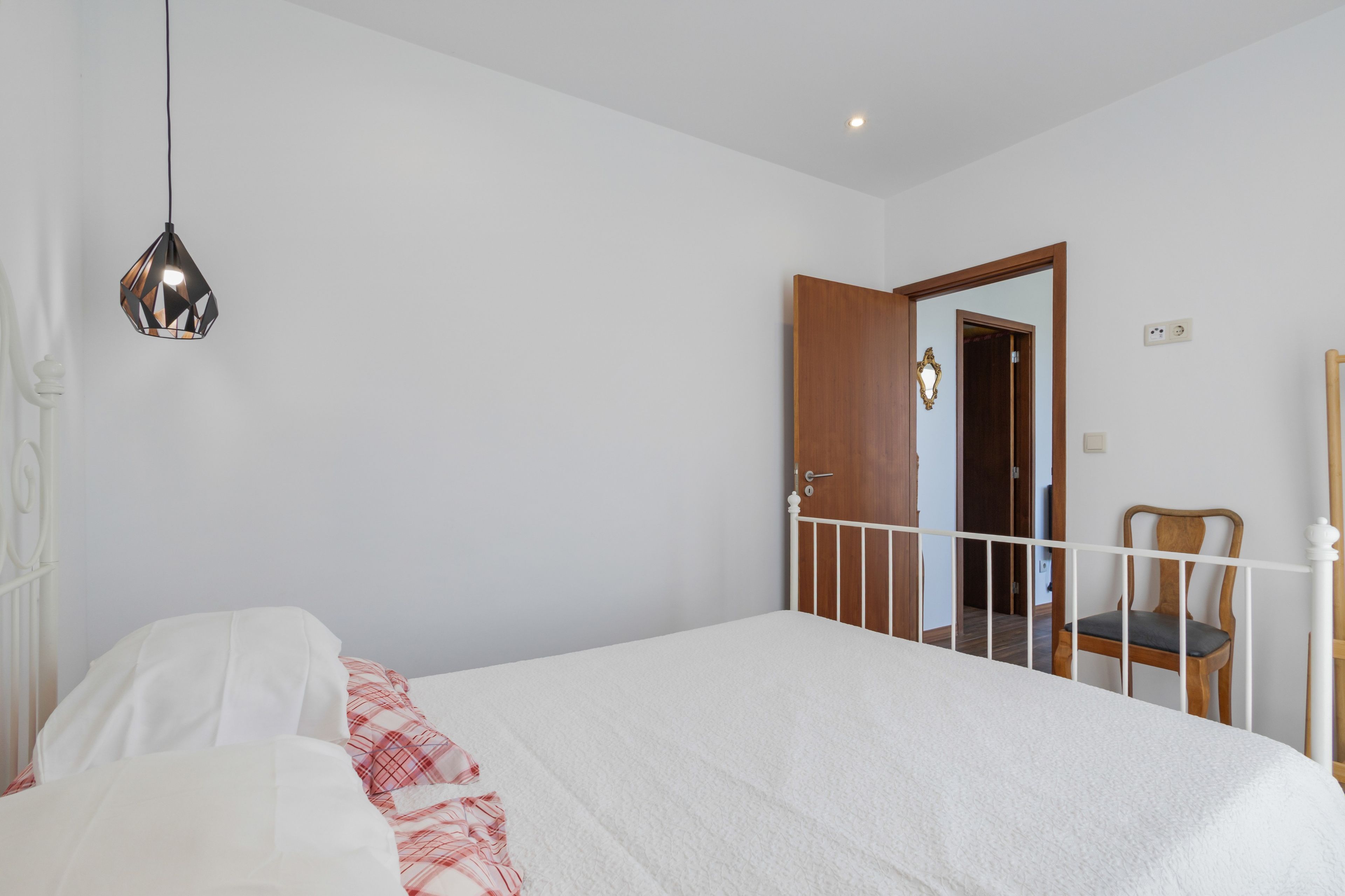 A bedroom with a bed, a bedside table, a chair, a hanging light fixture, and a door leading to another room.