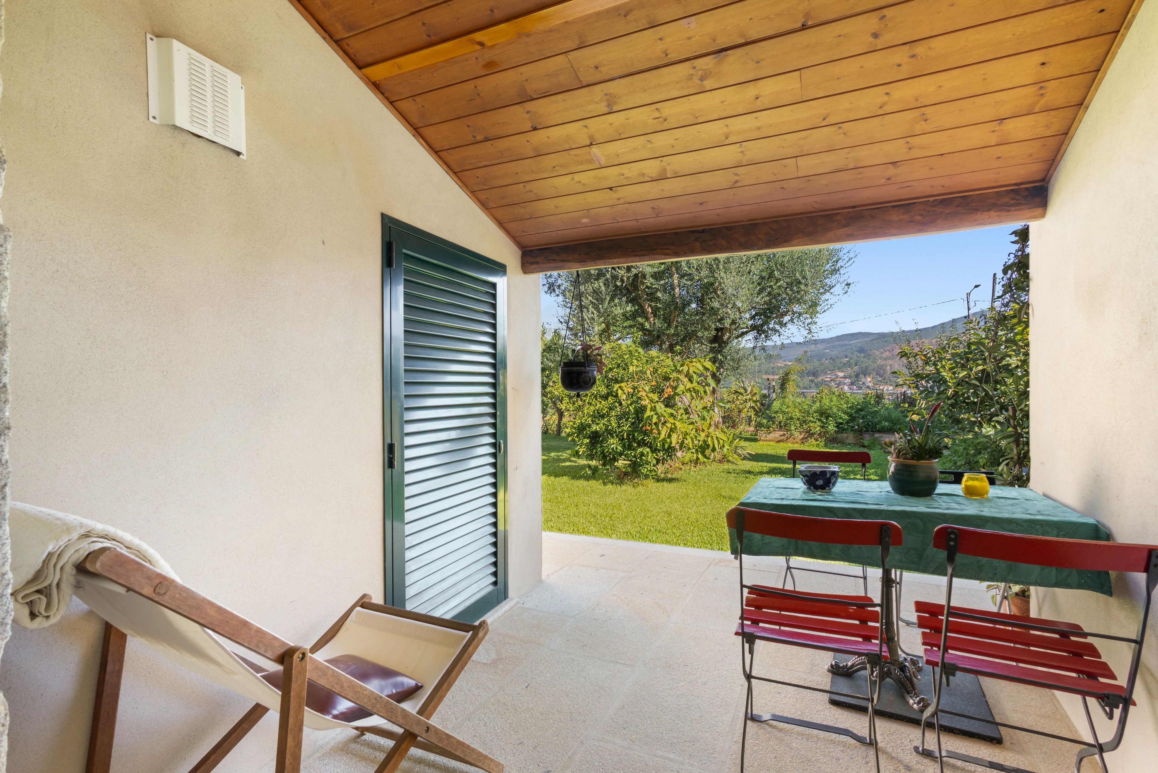 A covered outdoor seating area with a table and chairs, a door with shutters, and a view of a garden and hills in the background.