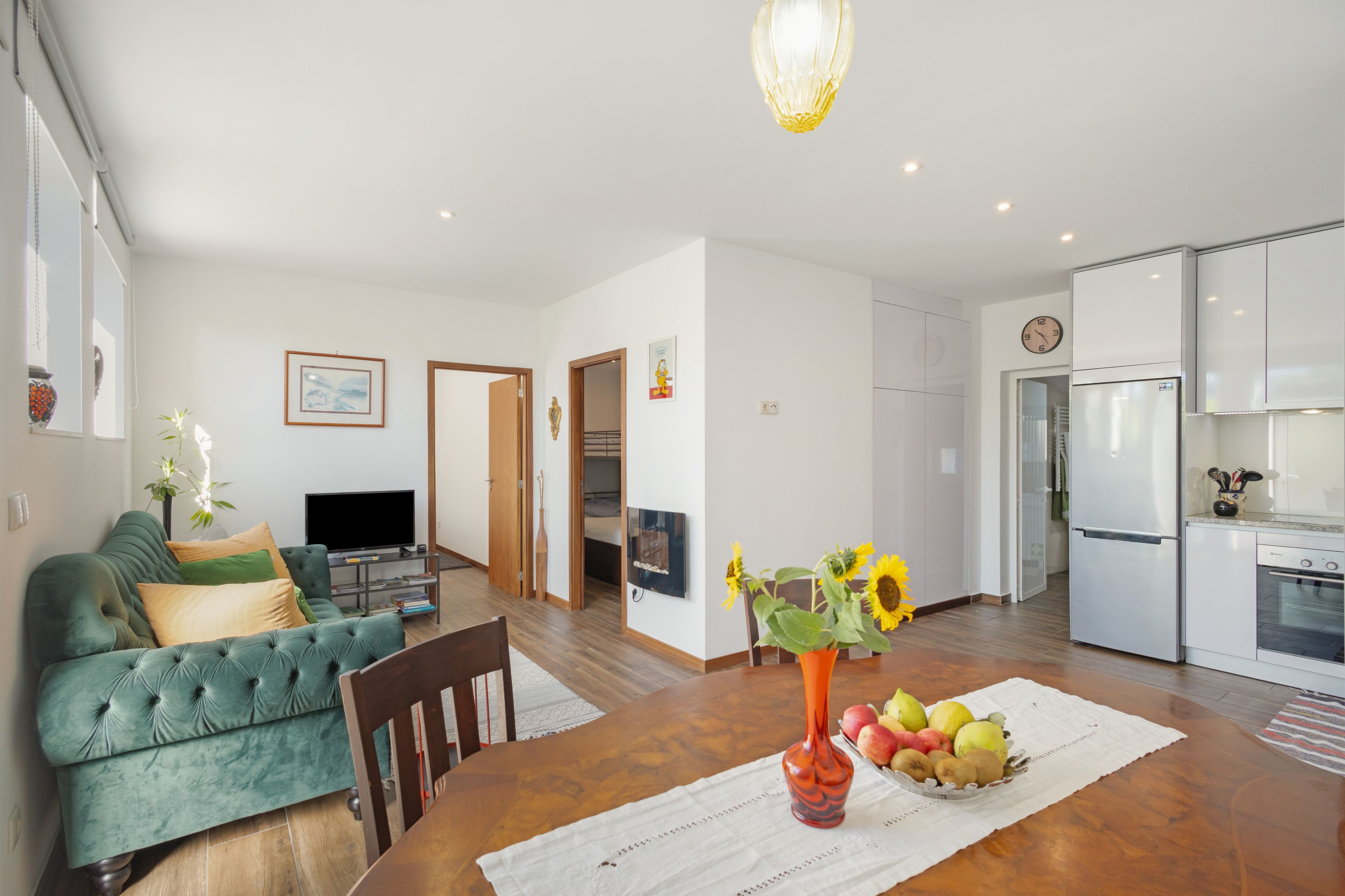A modern living room with a sofa, a dining table with a vase of flowers, and a kitchen area with a refrigerator.