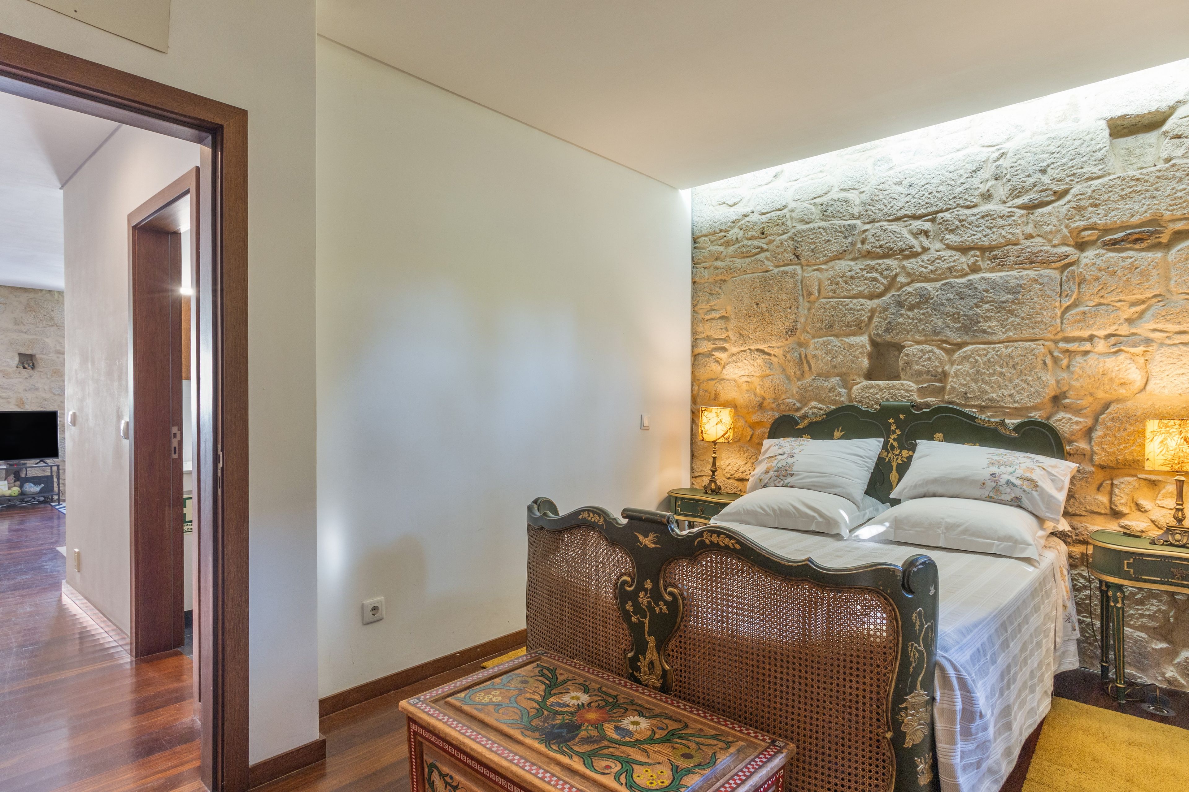A bedroom with a stone wall, a bed with white linens, a wooden chair, and a small table with a floral design.
