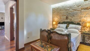 A bedroom with a stone wall, a bed with white linens, a wooden chair, and a small table with a floral design.