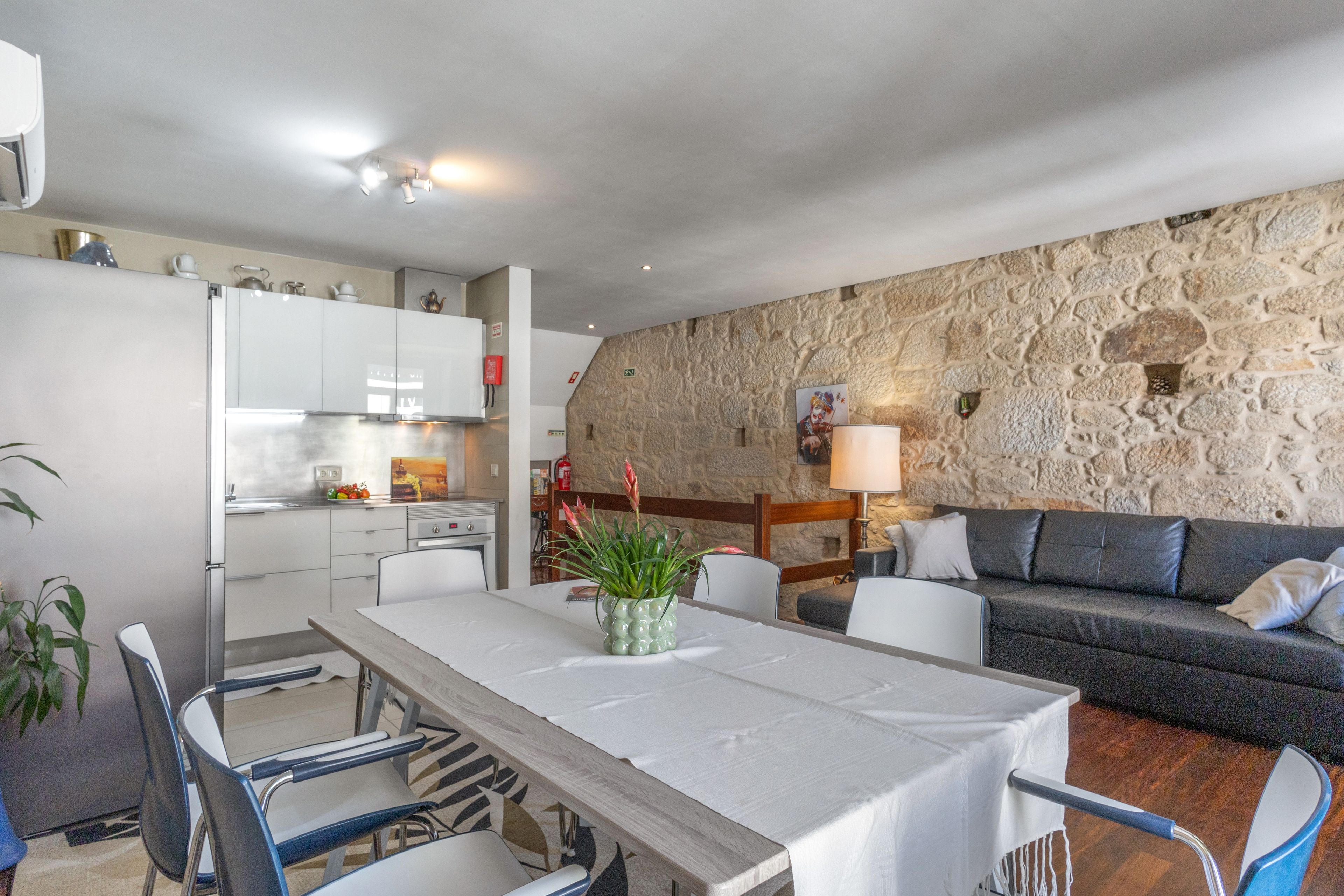 Eine moderne Küche mit weißen Schränken, einem Esstisch mit Stühlen, einer Steinwand und einem grauen Sofa in einem Wohnbereich.
