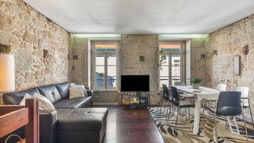 A living room with a stone wall, a leather sofa, a flat-screen TV, a dining table with chairs, and a patterned rug.