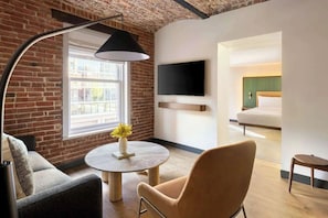A modern hotel room with a brick wall, a round table with a vase, a sofa, a television mounted on the wall, and a bed with a green headboard. - Junior Suite | Arlo DC | Downtown DC | Rooftop Bar (Washington)