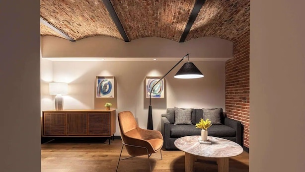 A room with a wooden floor, a round marble table, a leather armchair, a sofa, a cabinet, and a ceiling with exposed brick.