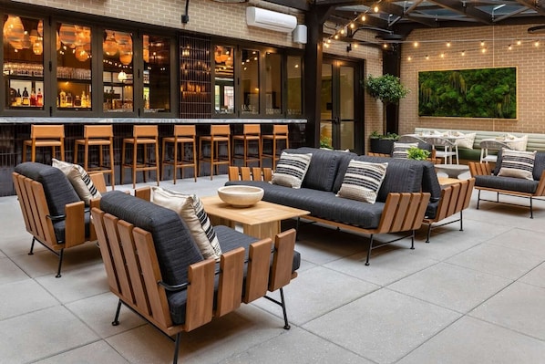 An outdoor seating area with sofas and chairs, a bar with bottles and glasses, and a large green wall.
