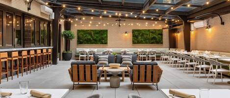 An outdoor dining area with multiple tables and chairs, a seating area with sofas, and a glass ceiling.