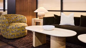 Un salon moderne avec un fauteuil jaune à motifs, un canapé foncé agrémenté d’un coussin blanc, une petite table d’appoint avec une lampe, et un bol blanc posé sur une table basse en bois.