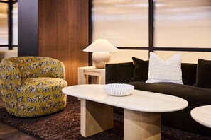 A modern living room with a yellow patterned armchair, a dark sofa with a white pillow, a small side table with a lamp, and a white bowl on a wooden coffee table.