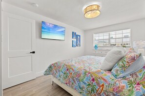 Ein Schlafzimmer mit einem Bett, einem Fenster mit Jalousien, einem an der Wand montierten Fernseher und einer Deckenleuchte.