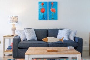 A living room with a dark grey sofa, patterned cushions, a wooden coffee table, and a wall-mounted lamp. - Coastal Stay by Dunedin Causeway (Dunedin)