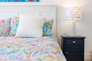 Ein Bett mit einem blumigem Überwurf, einem weißen Kopfkissen und einem Nachttisch mit einer Lampe.