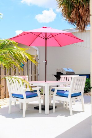 Eine Terrasse mit einem weißen Tisch und Stühlen, einem rosa Sonnenschirm und einem Grill.