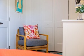 A room with a wooden chair, a colorful cushion, a white door, and a cabinet with a plant on top. - Coastal Dunedin Suite Near Beaches (Dunedin)