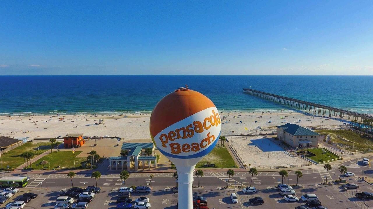 Udsigt over en strand med et stort orange og hvidt skilt der viser 'Pensacola Beach' og en mole, der strækker sig ud i vandet.