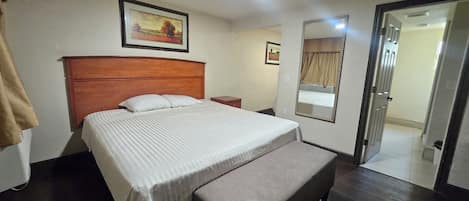 A hotel room with a bed, a bench, a nightstand, a mirror, and a door leading to another room.