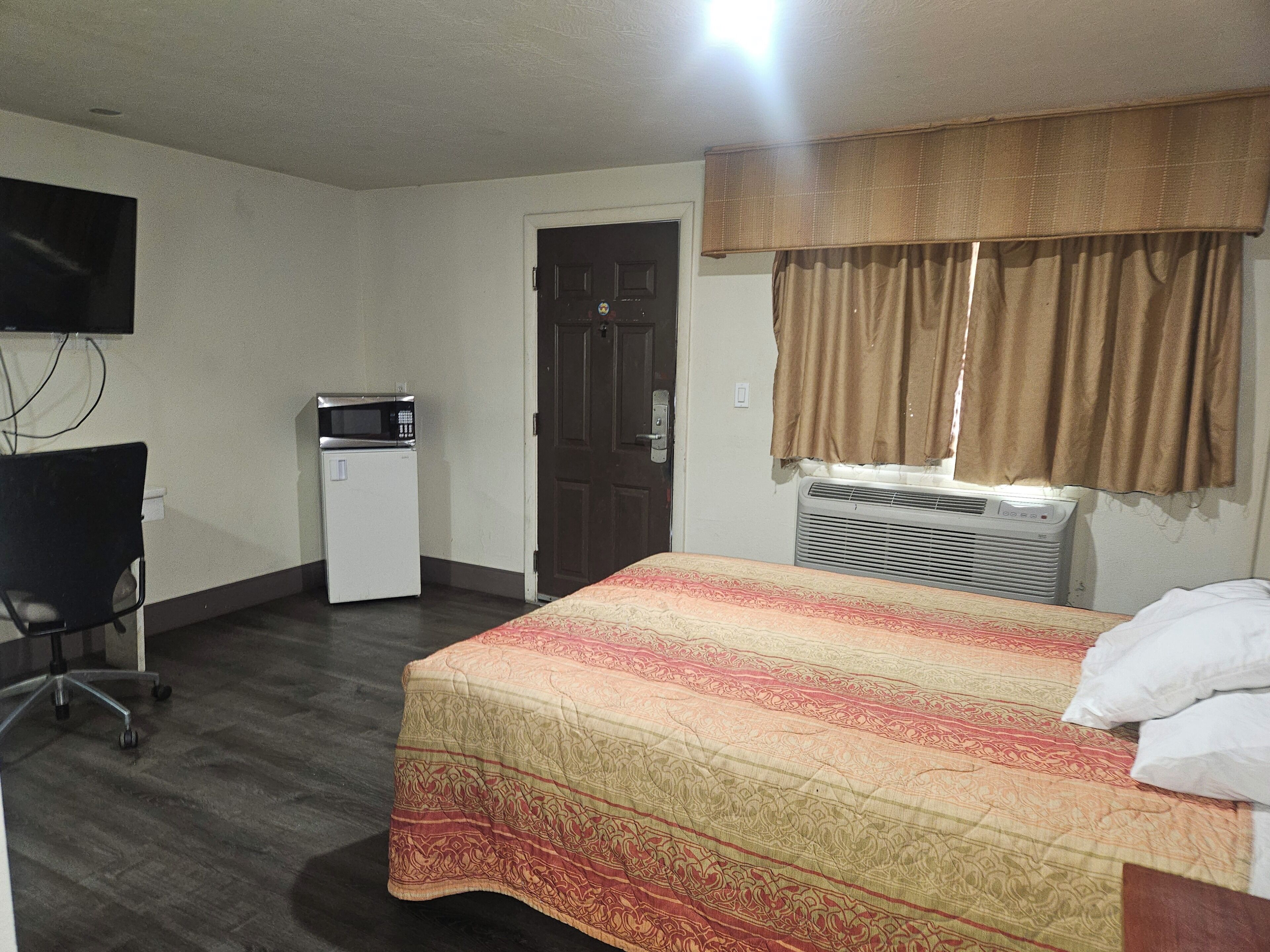 A hotel room with a bed, a desk, a chair, a microwave, a door, and a window with curtains.