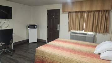 A hotel room with a bed, a desk, a chair, a microwave, a door, and a window with curtains.